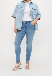 Kvinna som bär ljusblå denimjeans, vita klackar med remmar, vit figurnära topp och en lös ljusblå denimskjorta med nitar.