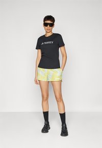 Juodas TERREX t-shirt'as, geltonos ir mėlynos raštuotos šortai, juodos kojinės ir juodi sportiniai bateliai. Apvalūs saulės akiniai, vidaus aplinka.