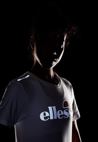 Hvid t-shirt med korte ærmer og reflekterende "ellesse" logo og emblem på brystet, der har en skinnende tekstur og glat stof.