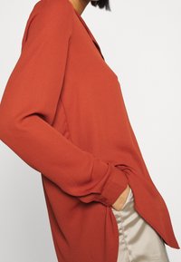 Blouse à manches longues couleur rouille avec un col en V, tissu lisse et fentes sur les côtés. Dispose d'un poignet à boutons et d'une coupe légèrement ample.