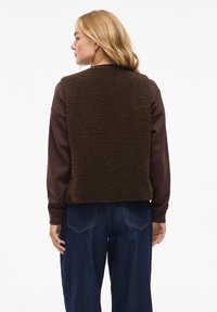 Braune Fleeceweste mit strukturierter Oberfläche, getragen über einem dunklen Langarmshirt. Sie hat einen abgerundeten Saum, weite Armlöcher und ist mit blauen Jeans kombiniert.