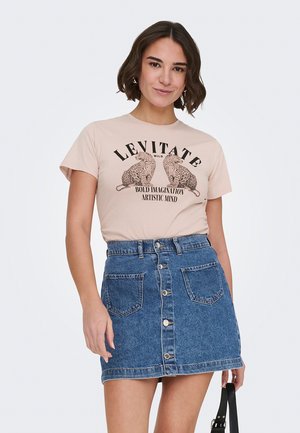 Frau trägt beige T-Shirt mit Leopardenmuster und dem Schriftzug "LEVITATE", kombiniert mit blauem Jeansrock mit Knöpfen, hält schwarze Umhängetasche.