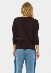 Femme aux longs cheveux blonds portant un haut ample marron foncé à manches trois-quarts et un jean bicolore bleu, debout de dos.