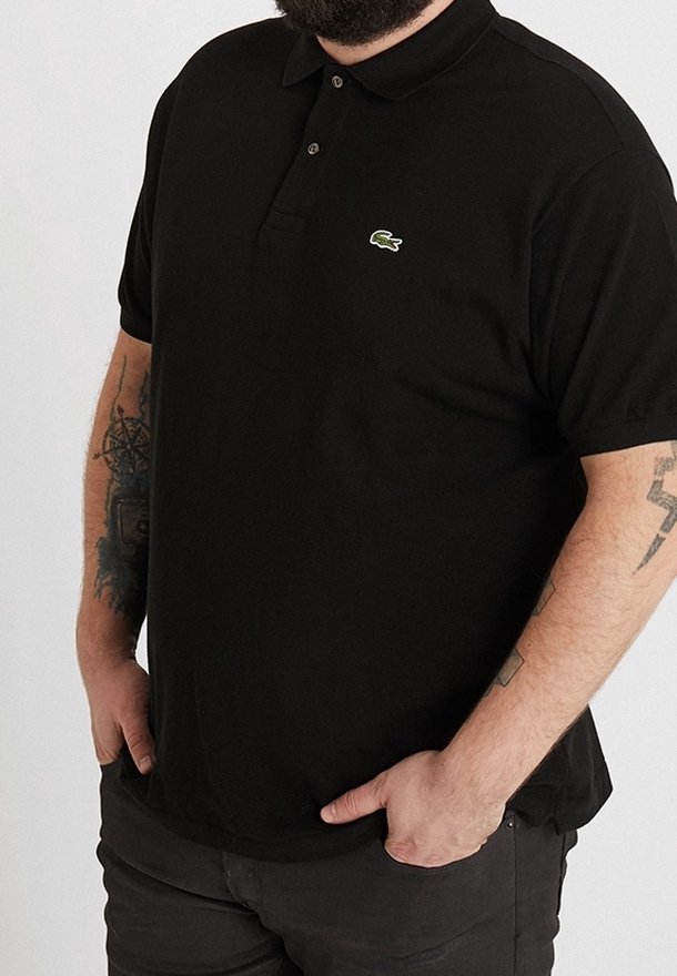 Homme portant un polo noir Lacoste avec le logo crocodile vert, pantalon noir, tatouages visibles sur les bras, mains dans les poches.