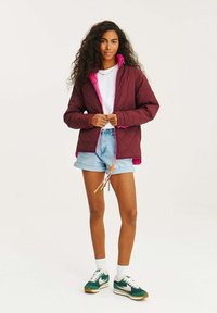 Bordeaux prošívaná bunda s růžovým vnitřkem a zipem, v kombinaci s světlými denimovými šortkami a zeleno-růžovými teniskami s bílými akcenty.