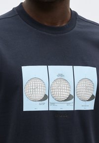 Άνδρας που φοράει σκούρο πουκάμισο με γραφικό που δείχνει τρία τεχνικά διαγράμματα της δομής και των μετρήσεων μιας μπάλας γκολφ.