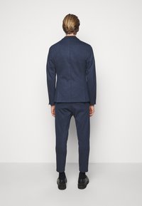 Completo blu navy in due pezzi realizzato in tessuto testurizzato, con giacca aderente e pantaloni slim, abbinato a scarpe nere. Vista da dietro.