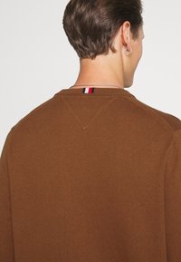 Maglione marrone lavorato a maglia con scollatura rotonda, caratterizzato da un piccolo accento a righe sul retro. La texture è liscia e uniforme.