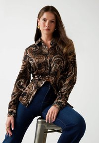 Donna con lunghi capelli, che indossa orecchini a cerchio dorati, una camicia bottonata marrone e nera con motivo paisley, jeans blu scuro, seduta su uno sgabello di metallo.
