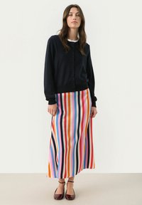 Cardigan blu navy con bottoni davanti, abbinato a una gonna maxi con strisce verticali multicolori. La modella indossa scarpe con cinturino alla caviglia color bordeaux.