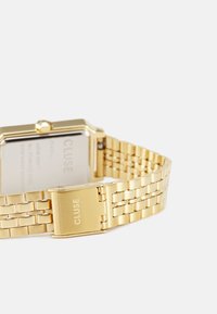 Montre-bracelet en métal doré avec un cadran rectangulaire et un bracelet à maillons. Dispose d'un fermoir avec le nom de la marque "CLUSE" gravé.