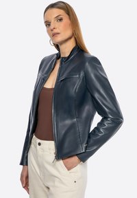 Marineblaue Leder-Motorradjacke mit Stehkragen, Reißverschluss und dezenten Fronttaschen. Trägt man über einem braunen Oberteil und hellfarbigen Hosen.