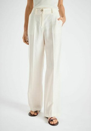 Vrouw draagt een crèmekleurige, wijde broek met hoge taille en plooien aan de voorkant, gecombineerd met donkerbruine teenslippers met ring en een mouwloos wit topje.