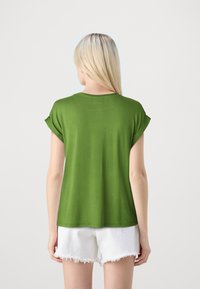 Grønn t-skjorte med korte ermer og rullede mansjetter, laget av myk stoff. Har rund hals og en avslappet passform. Brukt sammen med hvite slitte shorts.