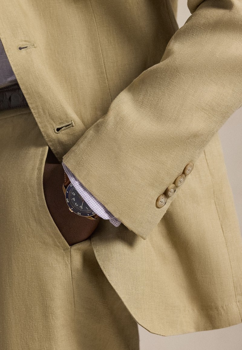 Beige linen suit with a notch lapel, featuring a pocket with a patterned shirt underneath. A watch peeks from the pocket.