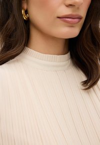 Top beige a collo alto plissettato con una texture liscia, caratterizzato da sottili linee verticali e un collo arrotondato. Orecchino a cerchio dorato visibile.