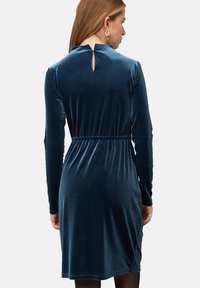 Samtkleid in tiefblau, mit langen Ärmeln, einem gerafften Bund und einem kleinen Schlüssel-Ausschnitt hinten. Glatte Textur und figurbetontes Design.
