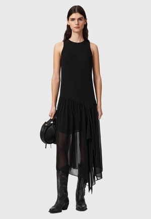 Kobieta w czarnej sukience bez rękawów z asymetryczną przezroczystą spódnicą, czarnych kowbojskich butach i trzymająca okrągłą czarną torebkę na szarym tle.