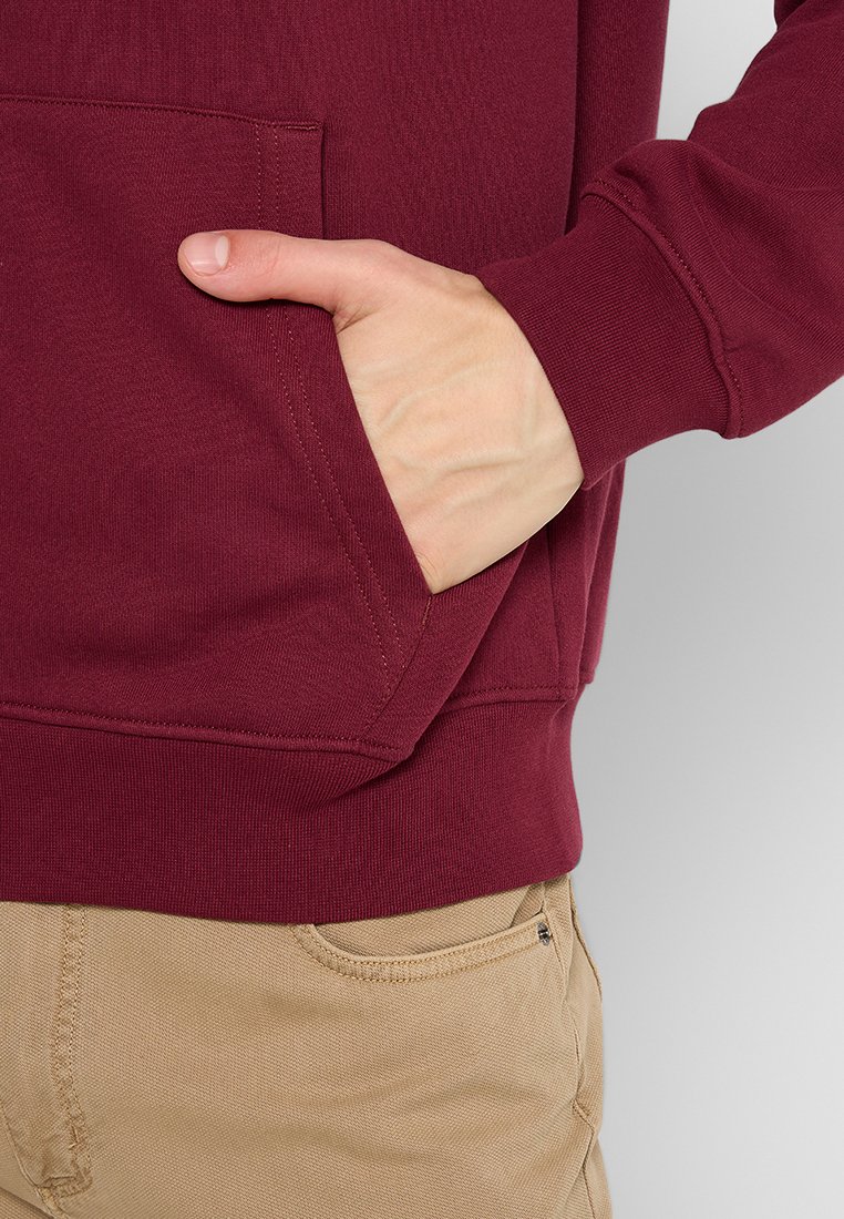 Hoodie bordeaux avec une poche kangourou. Le matériau est en coton texturé, avec un ourlet et des poignets côtelés. Les mains sont placées dans la poche.
