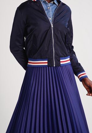 Blouson bomber bleu marine avec poignets et ceinture rayés rouge, blanc et bleu sur une chemise en jean, accompagné d'une jupe plissée bleu marine.