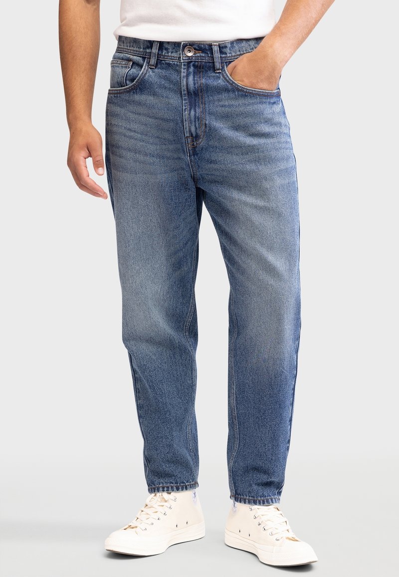 Homme portant un jean en denim bleu et des baskets blanches, debout avec une main dans la poche sur un fond uni.
