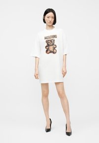 Γυναίκα με ίσια μαύρα μαλλιά, φοράει λευκό φαρδύ φόρεμα με αρκουδάκι και το κείμενο "MOSCHINO", σε συνδυασμό με μαύρες μυτερές γόβες.