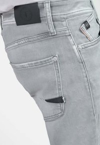 Grijze denim jeans met een gestructureerde afwerking, voorzien van meerdere zakken, een zwart leren patch bij de taille en knoopdetails op de zakken.