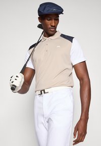 Polo bege com detalhes em azul-marinho e branco, calças brancas e um boné azul-marinho. Segurando um taco de golfe, usando uma luva de golfe branca.