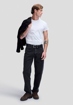 Mand iført hvid t-shirt, sorte jeans og brune sko, holder en sort jakke over skulderen og kigger til siden.