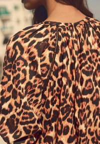 Blůza s leopardím vzorem a plisovanými detaily na zádech, v odstínech oranžové, černé a krémové. Měkká látka s viditelnými švy.