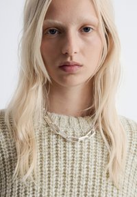 Silberne Kettenhalskette mit länglichen Gliedern, getragen über einem hellbeigen Strickpullover mit strukturiertem, geripptem Muster.