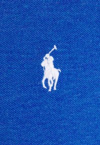 Logo de joueur de polo brodé blanc sur fond en tissu bleu texturé.