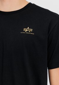 Černé bavlněné tričko s kulatým výstřihem a krátkými rukávy. Na levé části hrudníku se nachází zlaté logo. Měkká textura.