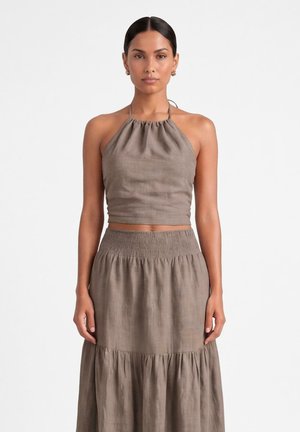 Kobieta o ciemnych włosach, ubrana w bezrękawnikowy crop top w kolorze taupe z dekoltem halter oraz pasującą wysoką taliowaną, marszczoną spódnicę midi na białym tle.