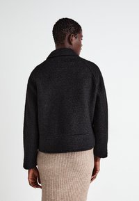 Black textured jacket with a collar and raglan sleeves, cropped length. Worn over a beige ribbed skirt, showcasing a simple design.