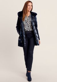 Donkerblauwe pufferjas met een nepbontkraag, ritssluiting zakken en een gequilt ontwerp, gecombineerd met een gebloemde blouse en donkere jeans.