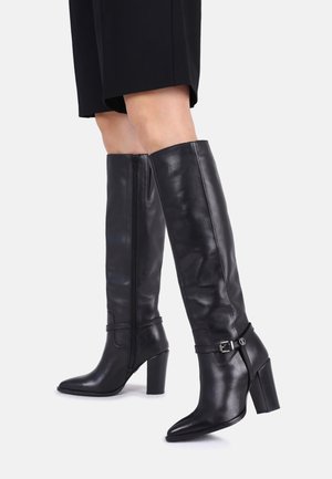 Personne portant des bottes en cuir noires montantes avec talons blocs et brides décoratives à boucle, debout jambes croisées.