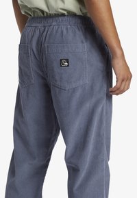 Blue corduroy pants with an elastic waistband and two back pockets, featuring a small black logo patch on the right pocket.