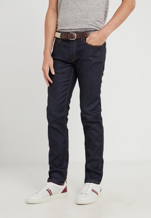 Donkerblauwe denim jeans met een slim fit, met oranje stiksels, vijfzakken ontwerp en een bruine riem met een gestructureerde afwerking. Sneakers met rode accenten.