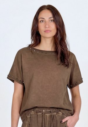 Žena s hnedými vlnitými vlasmi, ktorá nosí hnedý krátky top a zladené nohavice s malými kovovými cvokmi na okrajoch.