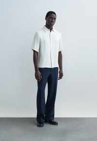 Άνδρας που στέκεται φορώντας ένα μπεζ πουκάμισο με κοντά μανίκια και κουμπιά, ναυτικού χρώματος παντελόνια με φαρδιά μπατζάκια και μαύρα παπούτσια μπροστά σε απλό γκρι φόντο.