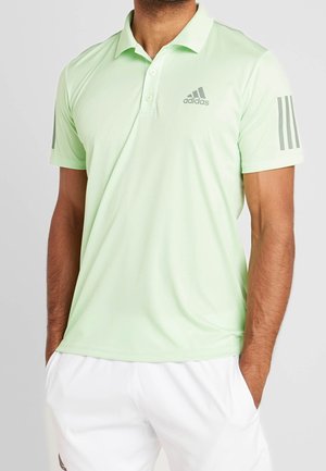 Homme portant un polo Adidas vert clair avec un logo gris et des bandes sur les manches, assorti à un short blanc, les mains dans les poches.