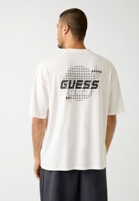 Biała koszulka z krótkim rękawem z czarnym wzorem w kratkę i logo "GUESS" na plecach. Wykonana z miękkiego, oddychającego materiału.