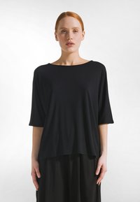 Roodharige vrouw met een bleke huid, gekleed in een losse zwarte top met mouwen tot aan de ellebogen en een zwarte broek, staand tegen een witte achtergrond.