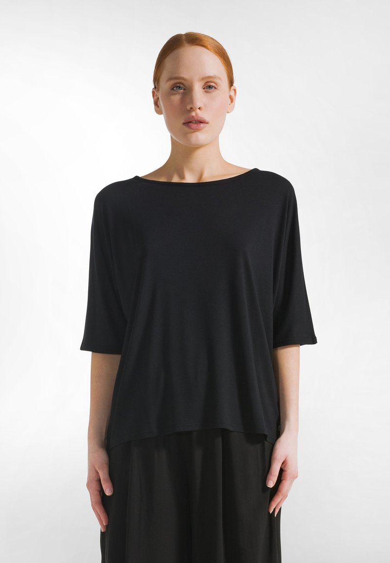 Roodharige vrouw met een bleke huid, gekleed in een losse zwarte top met mouwen tot aan de ellebogen en een zwarte broek, staand tegen een witte achtergrond.