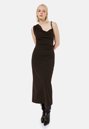 Lyshåret kvinde iført en sort ærmeløs midi-kjole med en strop og sorte spidse støvler, stående med hænderne bag ryggen.