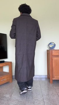 Casaco longo, castanho escuro, feito de um tecido macio, com gola alta, fenda nas costas e mangas longas; usado por cima de jeans pretos e ténis.