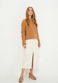 Suéter camel de punto trenzado y texturizado; falda crema con abertura frontal; zapatos taupe tipo slip-on con suelas elevadas. La modelo se encuentra de pie contra un fondo blanco.