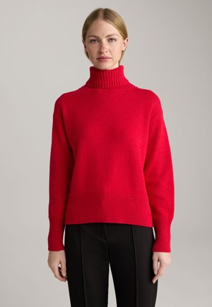 Rot gestrickter Rollkragenpullover mit gerippten Akzenten und entspannter Passform, kombiniert mit schwarzen, figurbetonten Hosen aus weichem, strukturiertem Material.