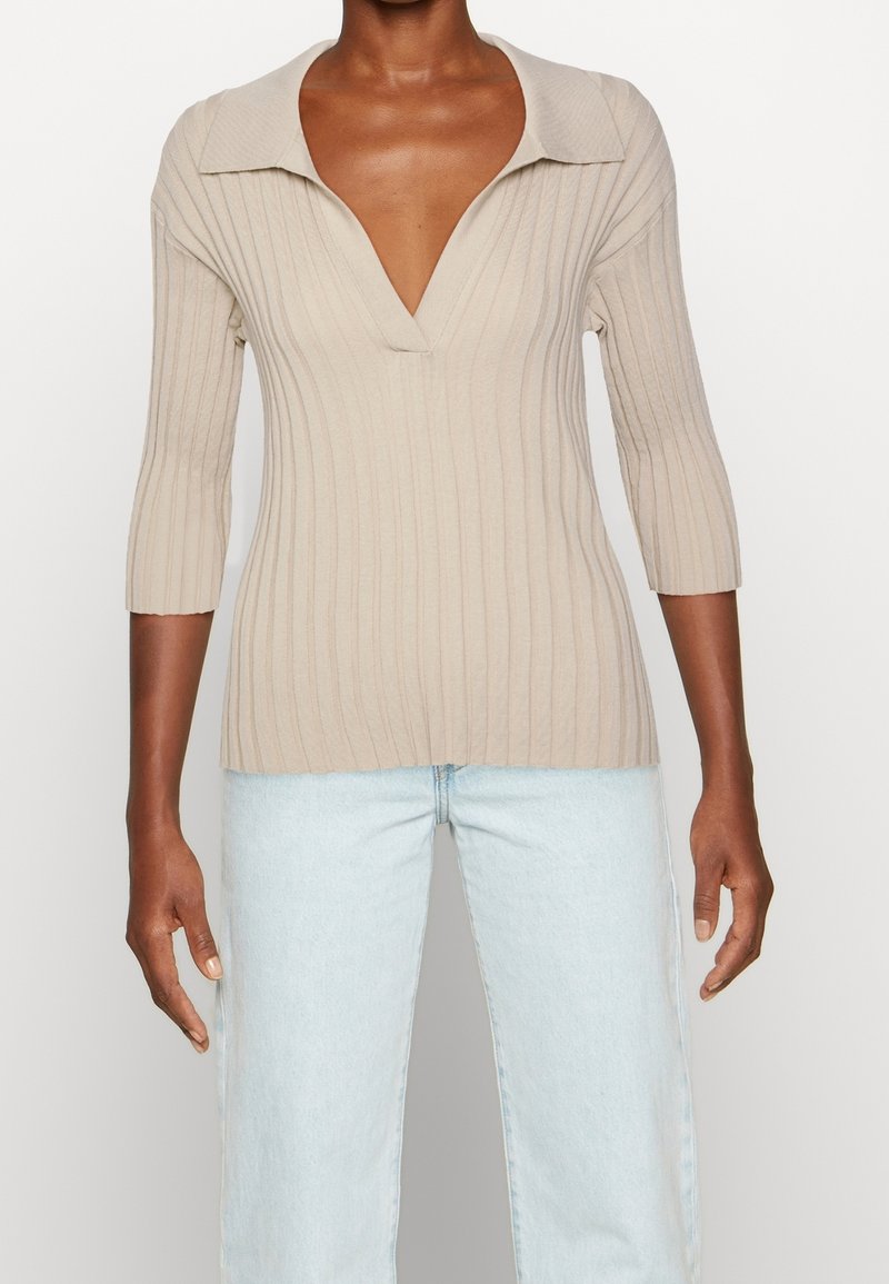 Personne à la peau foncée portant un pull beige côtelé à col en V avec manches 3/4 et un jean taille haute bleu clair sur fond blanc.